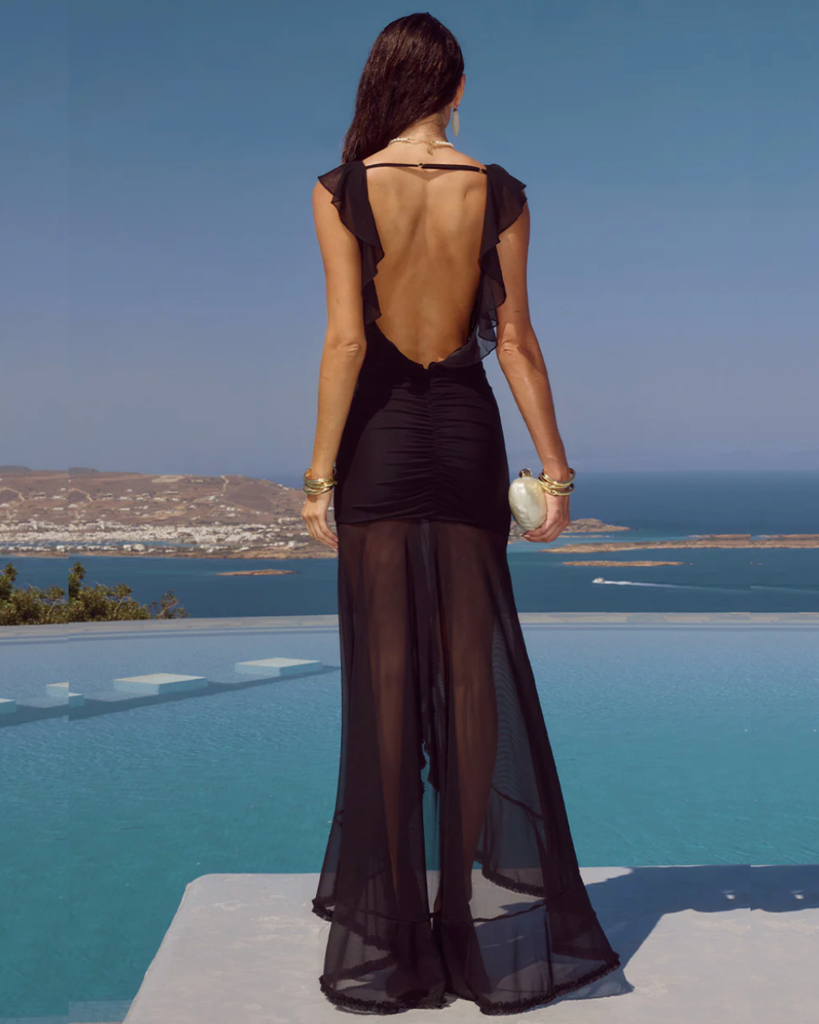 Woman in a black dress with sheer overlay standing by a pool with ocean view